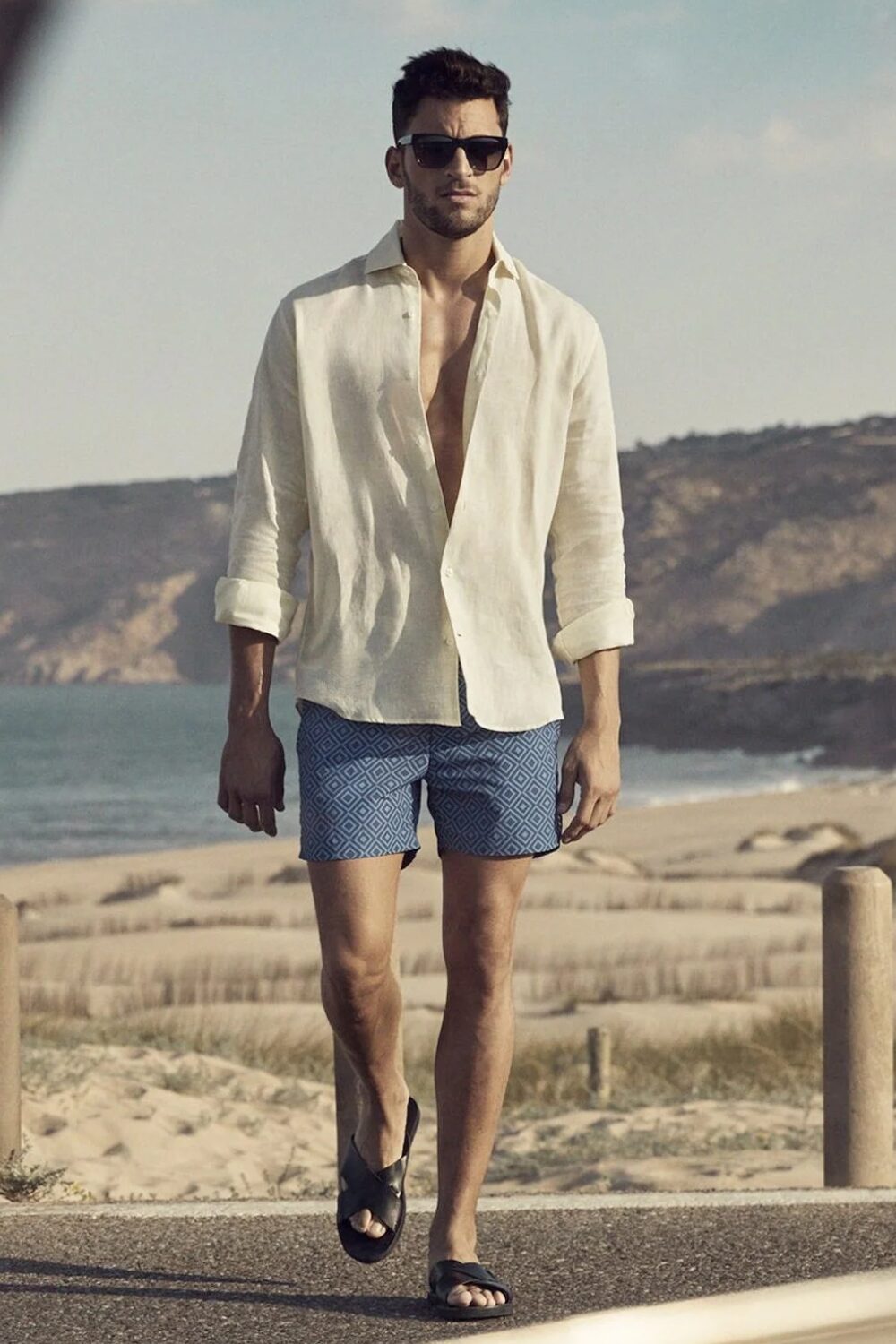 A man wearing swim shorts, a breathable linen shirt, comfortable sandals, and sunglasses — a perfect casual summer outfit for a beach day.